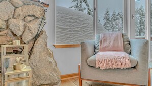 Tunnelmallinen huone, jossa on kiviseinä, puulattia, harmaa nojatuoli, jossa on vaaleanpunainen heittopeitto, sekä hylly, jossa on kynttilöitä ja metallinen kuppi.