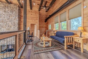 A wooden cabin interior with a stone wall, a ceiling fan, a sofa, a coffee table, and a window with a view of trees. - Cabin with Breathtaking Views, Hot Tub & Game Room (Sevierville)