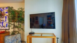 En stue med et vægmonteret fladskærms-tv, et træ tv-skab, en lænestol, en stor stueplante og et maleri på væggen.