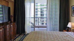 Et hotelværelse med seng, kommode, fjernsyn og balkon med udsigt til bygninger.