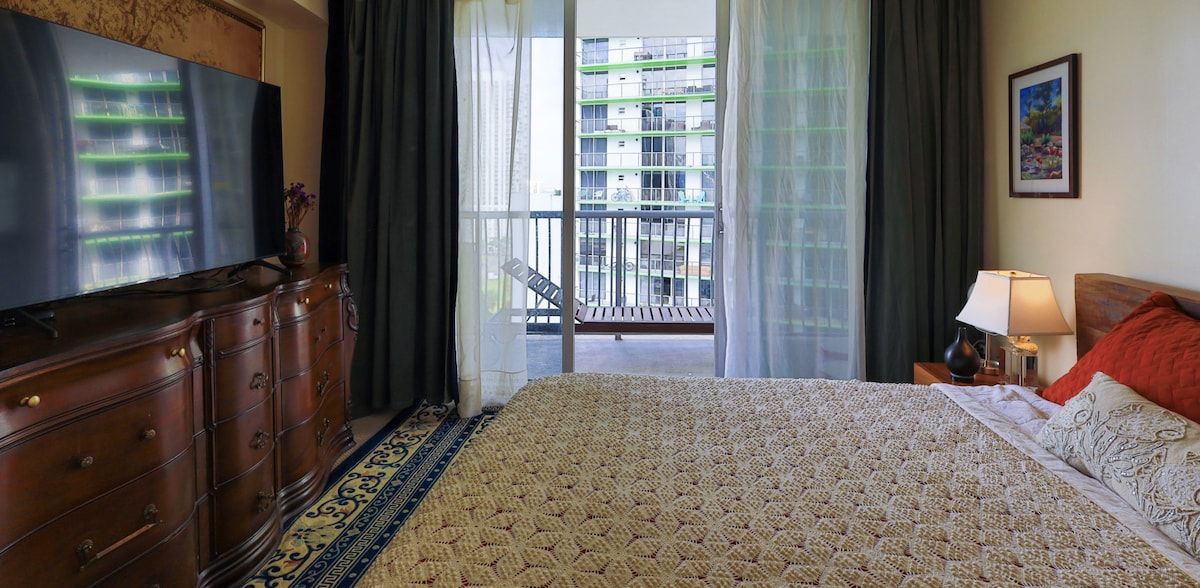 Quarto de hotel com uma cama grande, cômoda, televisão, varanda com vista para edifícios e um quadro na parede.