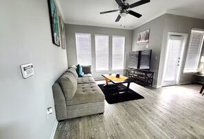 Ein Wohnzimmer mit grauem Sofa, Couchtisch, Deckenventilator und großen Fenstern mit Jalousien.