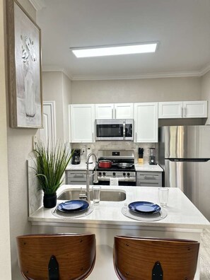 A modern kitchen with white cabinets, stainless steel appliances, a central island with seating, and a potted plant. - Chic 1BR Free Parking, Pool & Gym Near Woodlands (Magnolia)