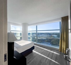 Ett hotellrum med ett stort fönster, en säng, ett sängbord och utsikt över stadsbilden.