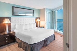 A hotel room with a large bed, two bedside tables, a mirror above the bed, and a view of the sea through a large window. - Oceans One 1008 (Myrtle Beach)