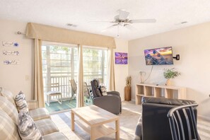 A spacious living area with a sofa, armchair, and coffee table, a television mounted on the wall, and a sliding glass door leading to an outdoor patio. - River Oaks 19-E (Myrtle Beach)