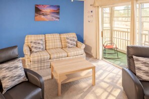 A living room with a sofa, two armchairs, a coffee table, a painting, and a sliding glass door leading to an outdoor area. - River Oaks 19-E (Myrtle Beach)