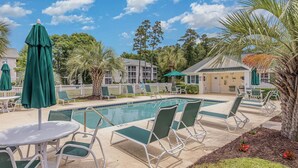 Área de piscina com espreguiçadeiras e guarda-sóis, rodeada por palmeiras e um edifício.