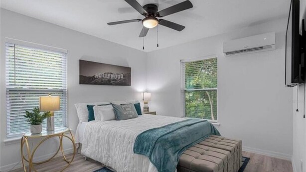 Une chambre à coucher comprenant un lit, un ventilateur de plafond, une table de chevet, une lampe, une fenêtre et un téléviseur fixé au mur.
