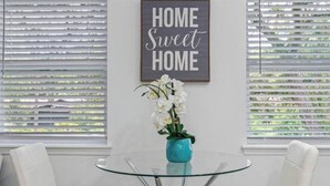Une pièce avec une table en verre, deux chaises blanches, un vase bleu avec des fleurs blanches, et une pancarte au-dessus sur laquelle on peut lire : « Chez soi, on est bien ».
