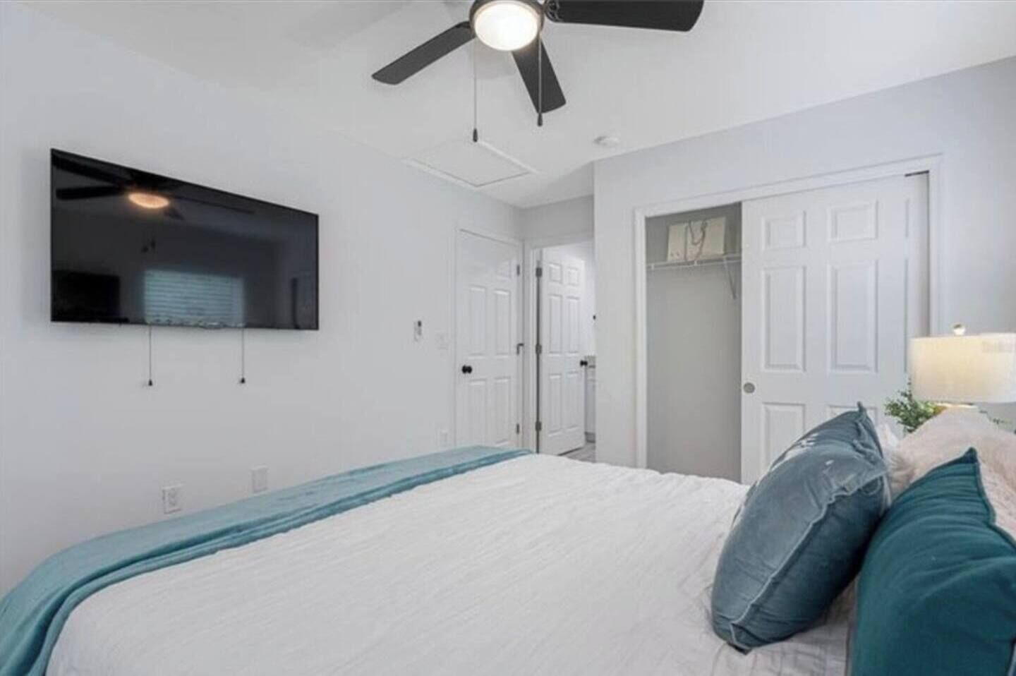 A bedroom with a bed, a ceiling fan, a TV mounted on the wall, and a door leading to another room.