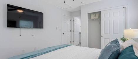 A bedroom with a bed, a ceiling fan, a TV mounted on the wall, and a door leading to another room.