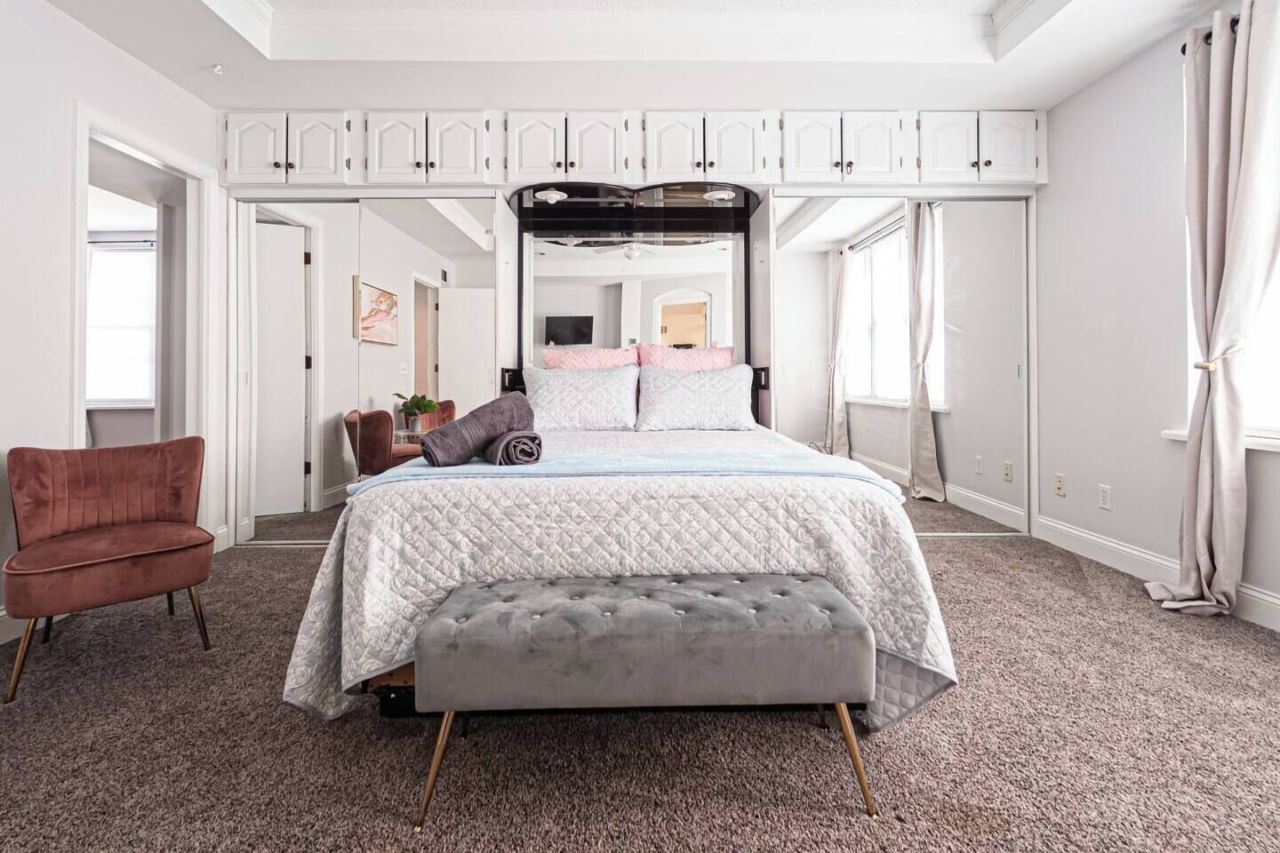 Une chambre à coucher avec un grand lit, un banc gris, une chaise rouge et des meubles blancs.