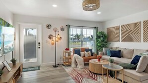 A living room with a sofa, coffee table, and floor lamp. There is a window with blinds, a door, and a wall with decorative panels. - Newly Remodeled Lush Boho Vibe (Apollo Beach)