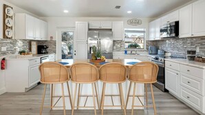 A modern kitchen with white cabinets, a stainless steel refrigerator, and a dining area with four wicker chairs. - Newly Remodeled Lush Boho Vibe (Apollo Beach)