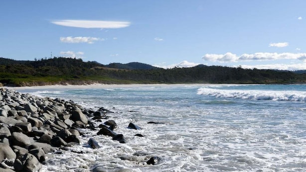 A rocky beach with waves, a clear sky, and distant hills.
