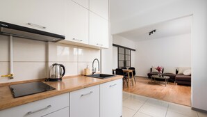 Una cocina moderna con armarios blancos, encimera de madera y una placa de inducción integrada. Hay una zona de comedor con mesa y sillas, y un sofá al fondo.