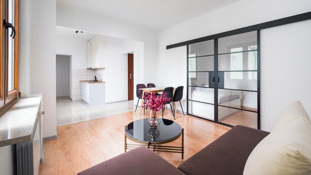 Una sala moderna con un sofá, una mesa de comedor con sillas, una cocina al fondo y un ventanal corredero que da a un balcón.