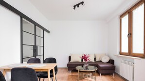 Una sala de estar moderna con una mesa de comedor de madera, sillas negras, una mesa de centro de vidrio con un florero, un sofá marrón con cojines y un ventanal con persianas.