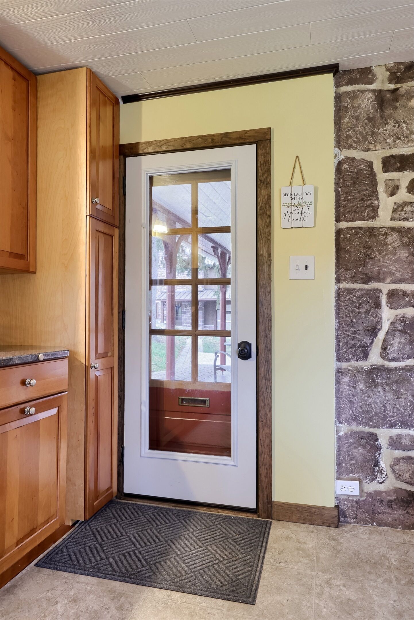 A kitchen with wooden cabinets, a door with a window, a stone wall, and a mat at the entrance.