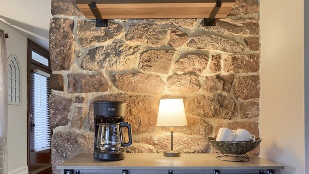 A stone wall with a shelf holding a coffee maker, a lamp, and a basket. Below is a wooden cabinet with two doors. - Cedar Glen Farmhouse • 1800s Stone Retreat (Mohnton)