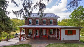 A two-story stone building with a covered porch, a brick pathway leading to the entrance, and a small white structure with red shutters to the right. - Cedar Glen Farmhouse • 1800s Stone Retreat (Mohnton)