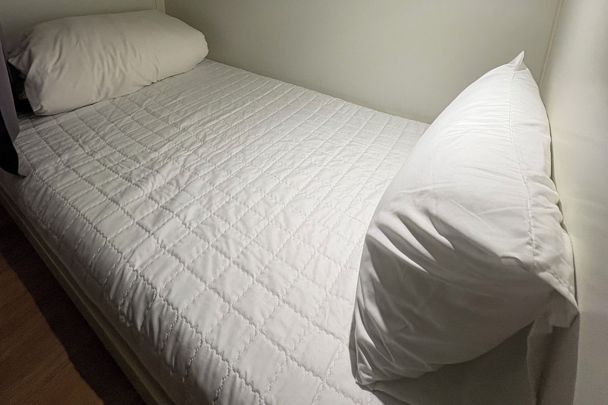 A small, single-bed room with a white quilted bedspread and pillows, a white headboard, and a ceiling light.