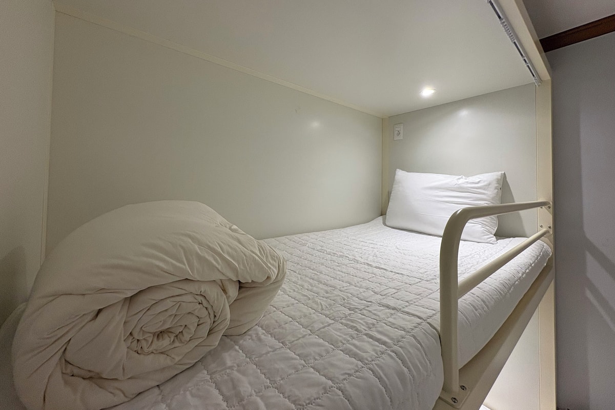 A small, single-bed room with a white bed, a rolled-up blanket, and a white wall.