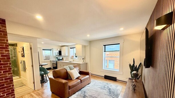 A spacious living area with a brown leather sofa, a wooden coffee table, and a large window providing natural light. A kitchen with white cabinets and a dining area with a green chair are visible in the background. - Modern Rustic Luxury - 1 Bdrm (Moncton)