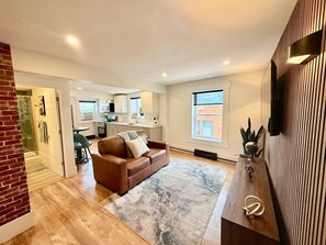 A spacious living area with a brown leather sofa, a wooden coffee table, and a large window providing natural light. A kitchen with white cabinets and a dining area with a green chair are visible in the background. - Modern Rustic Luxury - 1 Bdrm (Moncton)