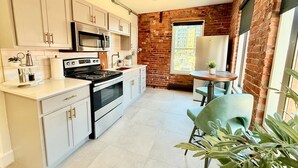 A modern kitchen with white cabinets, stainless steel appliances, and a brick wall. There is a dining area with a table and chairs. - Modern Rustic Luxury - Studio (Moncton)