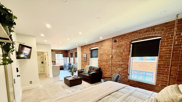 A modern bedroom with a large bed, a TV, and a brick wall. - Modern Rustic Luxury - Studio (Moncton)