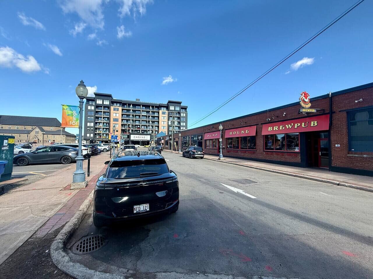 A street view with a black SUV parked on the side, a building with a red awning labeled "BREWPUB," and a multi-story building in the background.