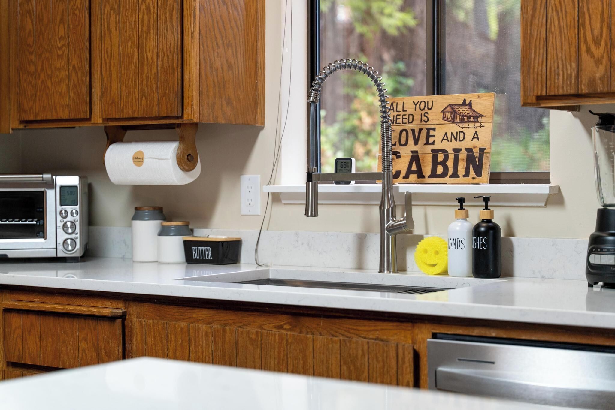 A kitchen with wooden cabinets, a stainless steel sink, a roll of paper towels, a toaster, and a sign above the sink.