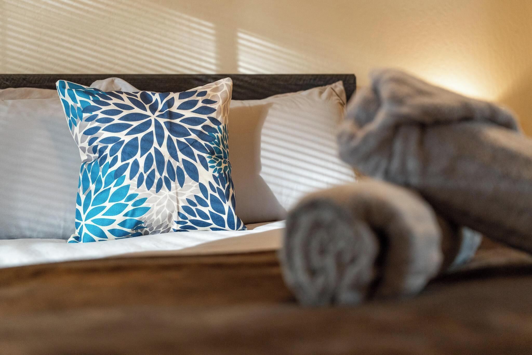 Un lit bien fait, avec un oreiller à motifs bleus et blancs, une serviette enroulée et une tête de lit en bois.