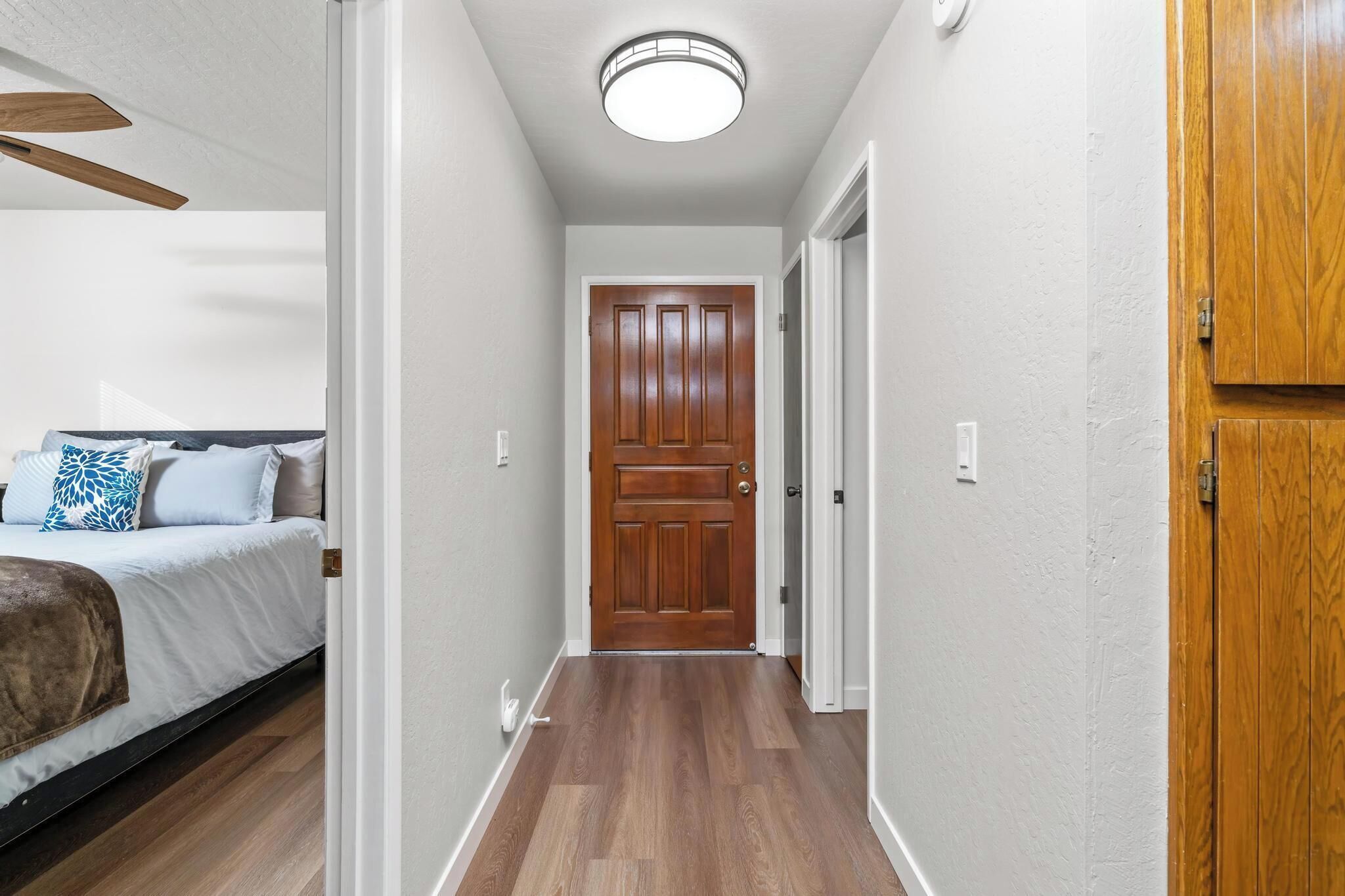 Un corridoio stretto con una porta di legno in fondo, un letto con cuscino bianco e azzurro, e una ventola a soffitto.