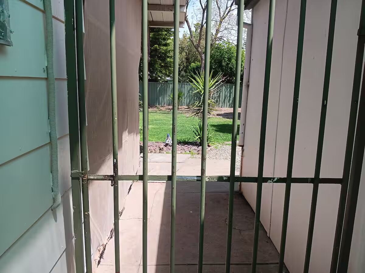 Un étroit couloir extérieur, avec une grille en métal, un sol en béton et une vue sur une cour verdoyante, délimitée par une clôture et agrémentée d’arbres.