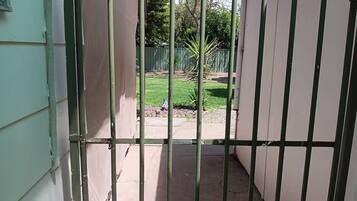 Un étroit couloir extérieur, avec une grille en métal, un sol en béton et une vue sur une cour verdoyante, délimitée par une clôture et agrémentée d’arbres.