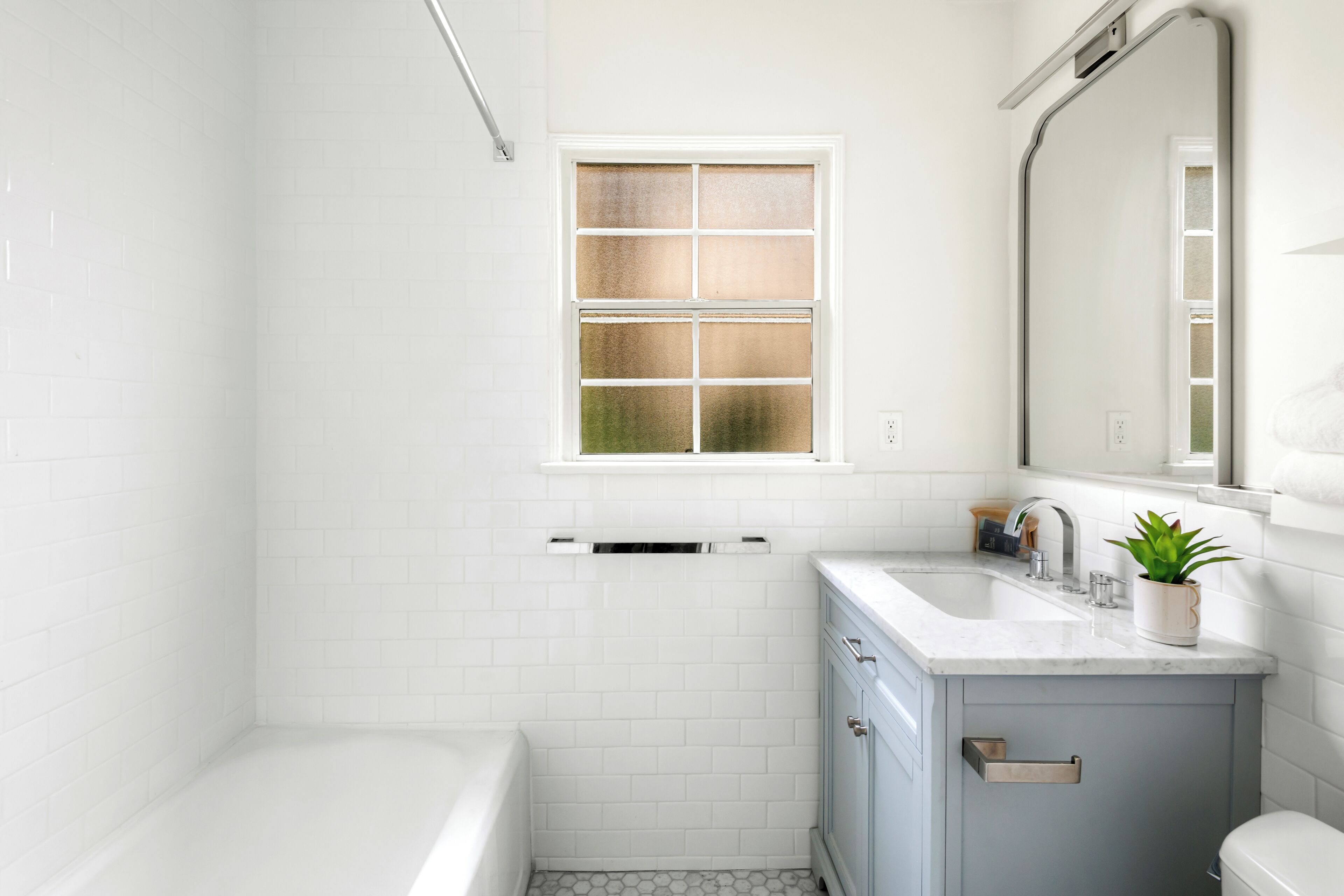 Ein Badezimmer mit weißen Fliesen, einem Fenster, einem Waschbecken, einem Spiegel und einer Toilette.