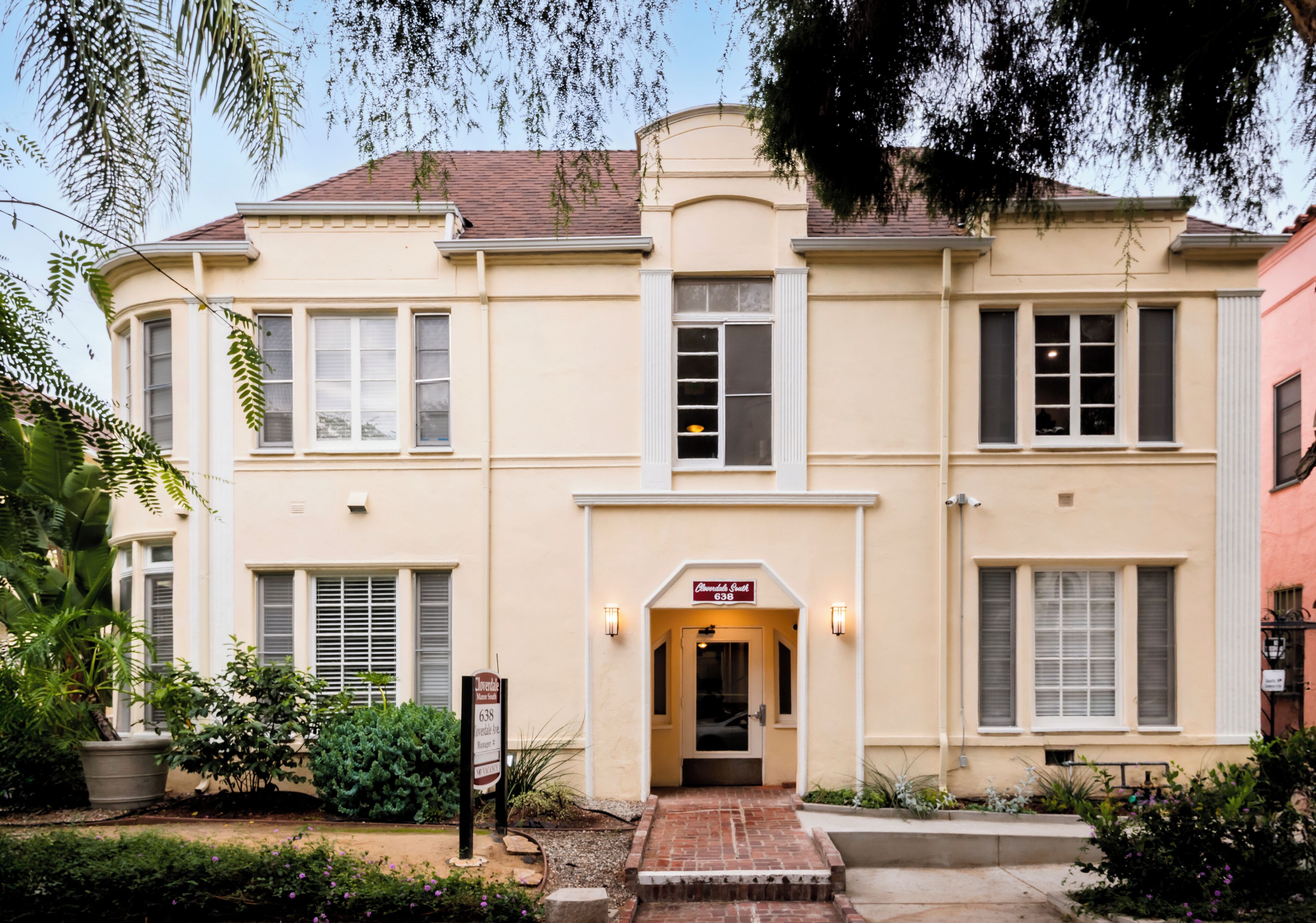 A two-story building with a beige facade, a central entrance with a brick pathway, and a sign above the door.
