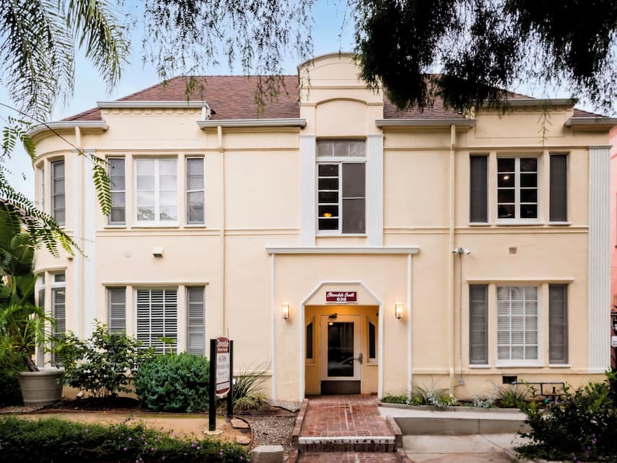 A two-story building with a beige facade, a central entrance with a brick pathway, and a sign above the door.