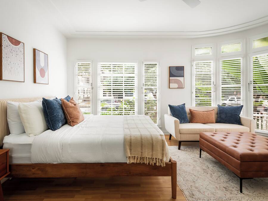 A bedroom with a bed, a sofa, a bench, and large windows with blinds.