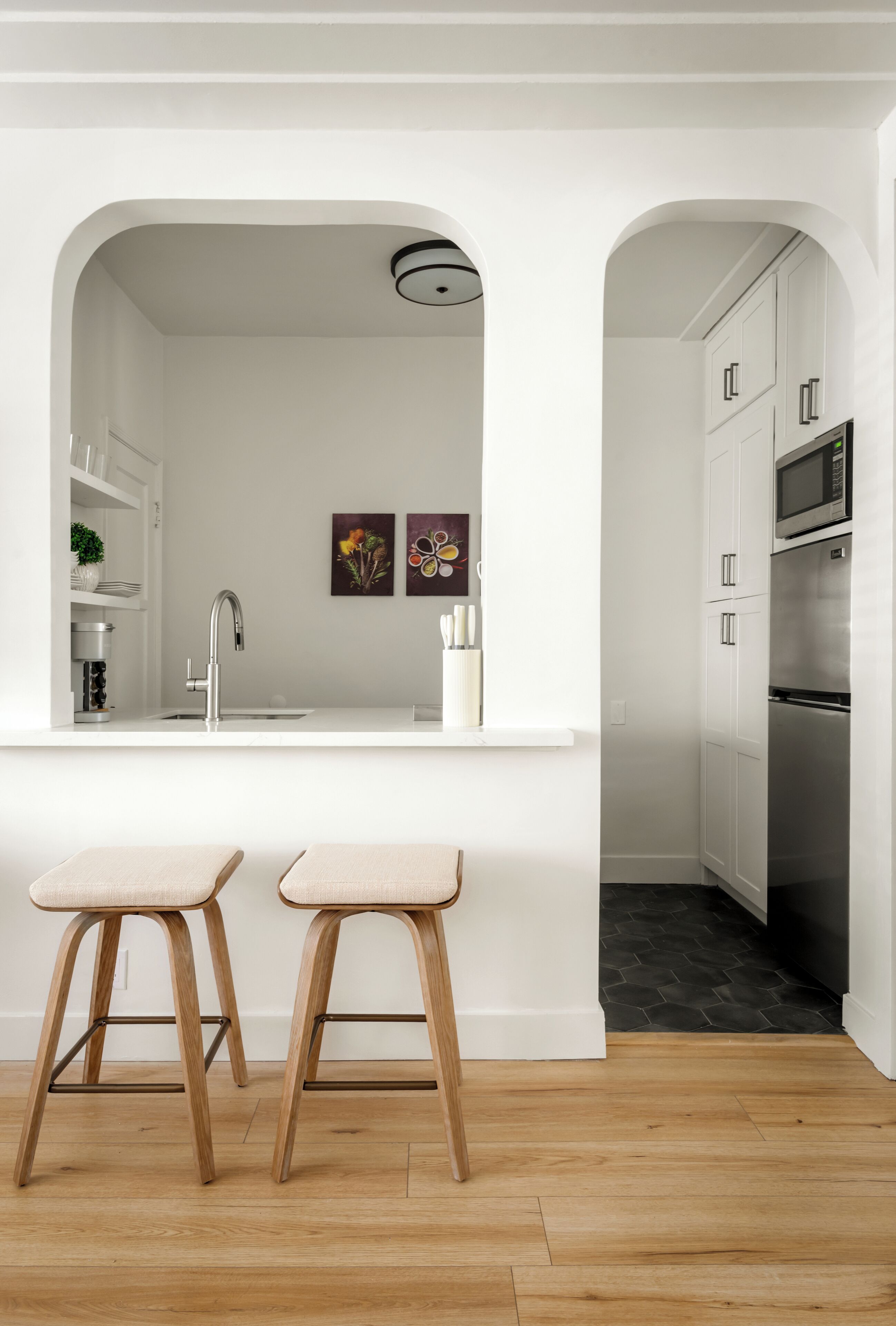 Una cocina moderna con armarios blancos, un refrigerador de acero inoxidable y una encimera blanca. Hay dos taburetes de madera y un lavamanos con grifo.