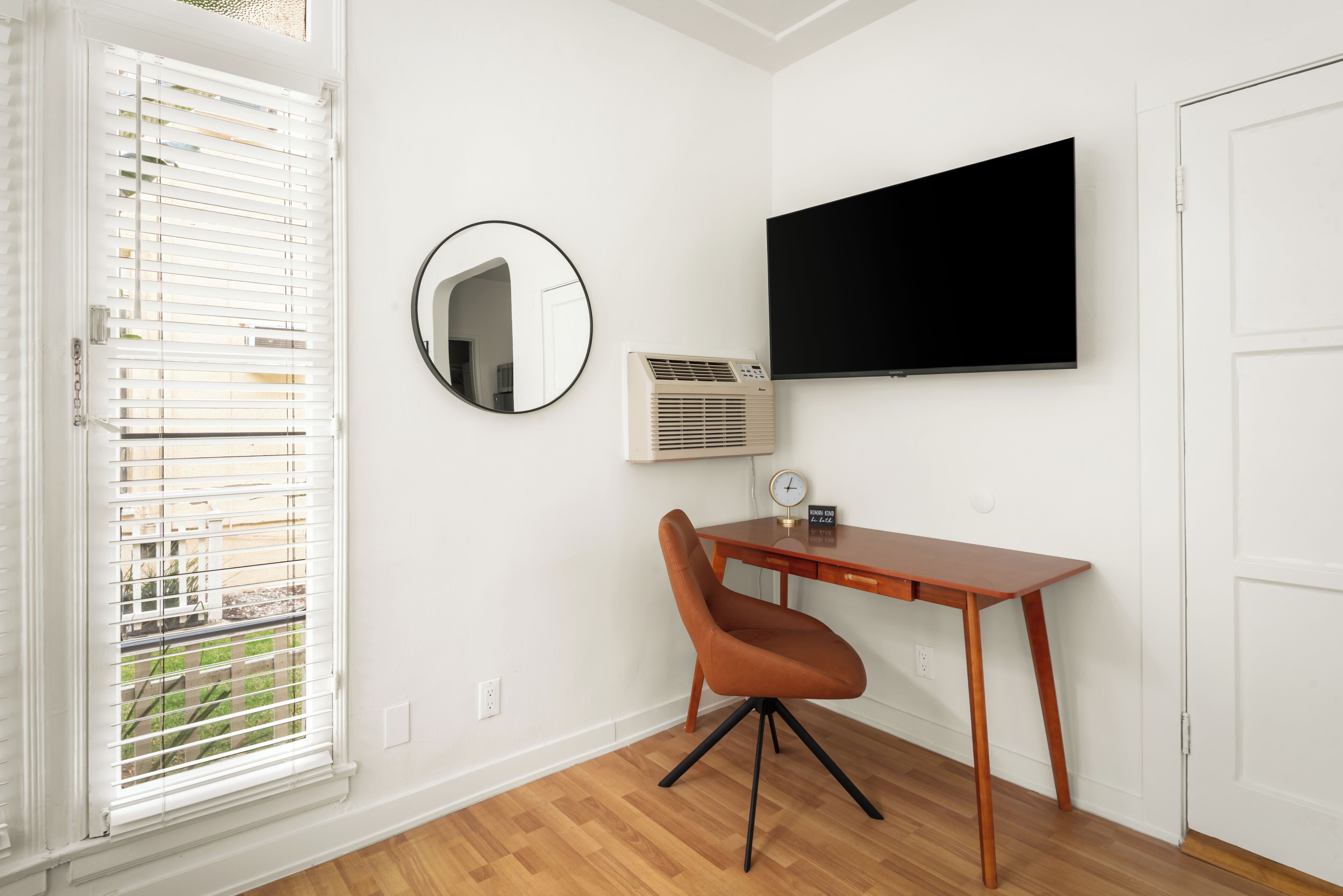 A room with a wooden desk, a chair, a mounted TV, a round mirror, and an air conditioning unit.