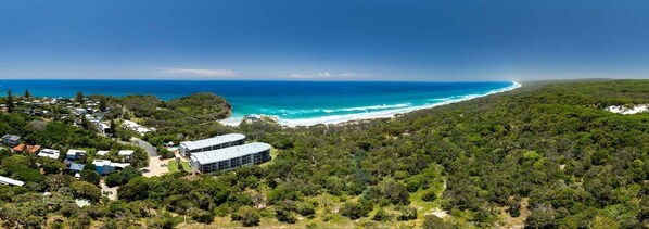 An aerial view of a coastal area with a beach, dense greenery, and a building with a modern design.
