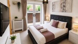 A bedroom with a bed, a TV, a desk with a remote, a radiator, and a window with curtains. - The Grounded Pearl (Salford)