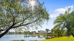 A serene lakeside view with a well-maintained lawn, a large tree, and a row of houses in the background. - Beautiful vacation home with great lake view and pool in Naples (Naples)
