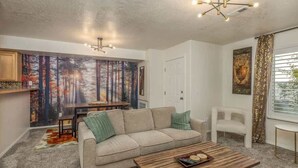 A living room with a beige sofa, a wooden coffee table, and a dining area with a table and chairs. There is a painting of a forest on the wall, a white door, and a window with curtains.