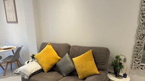 A modern living room with a grey sofa, yellow and grey cushions, a small side table with a plant, and a decorative mirror.