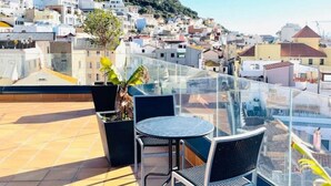 A rooftop terrace with a glass balustrade, a round table, two chairs, and a potted plant.